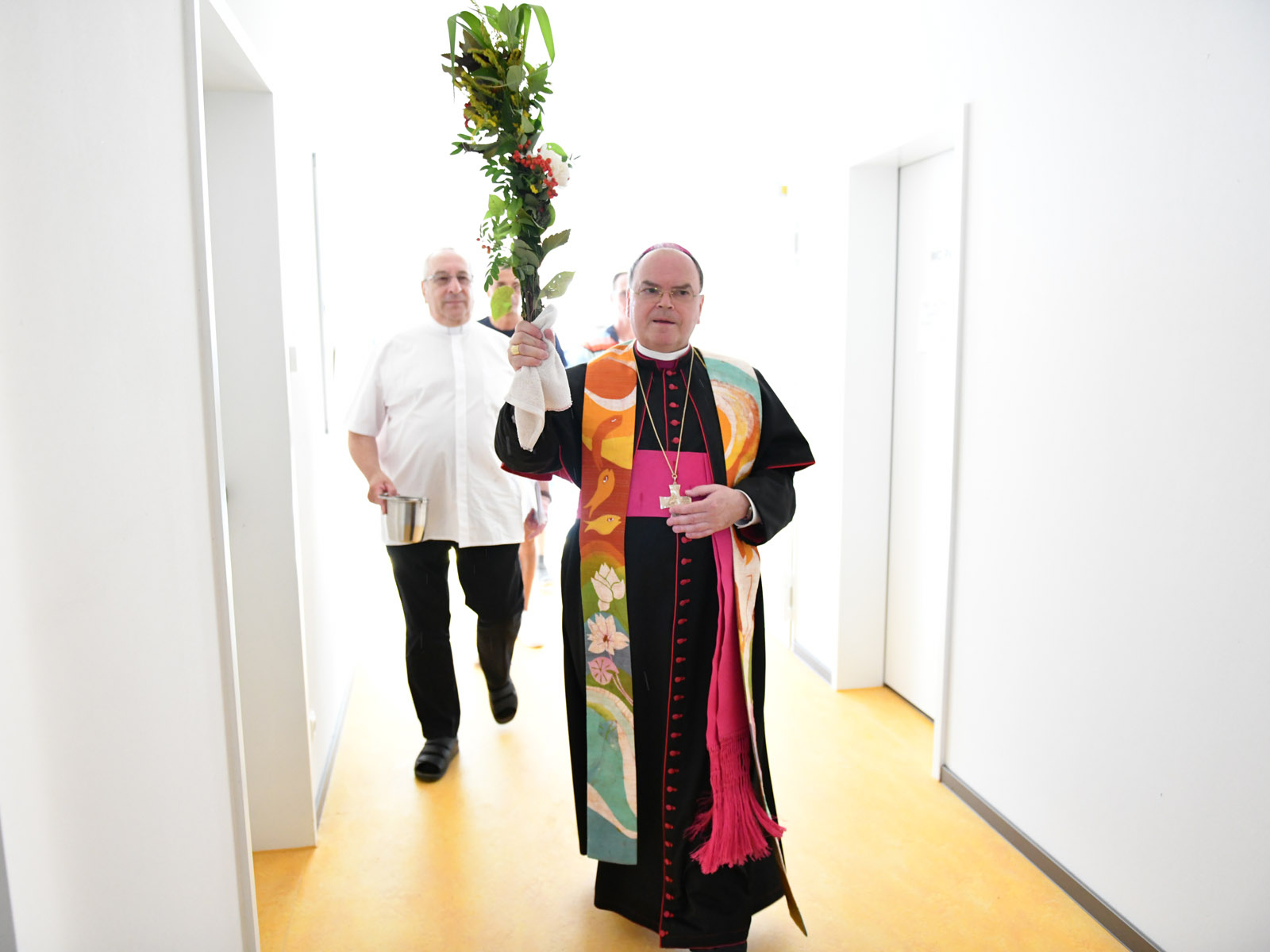 Der Bischof weiht das neue Wohnheim ein. Als Weihwasserpinsel verwendete er dabei einen Büschel mit Blumen und Gräsern aus dem Garten des Abbé-Pierre-Zentrums.